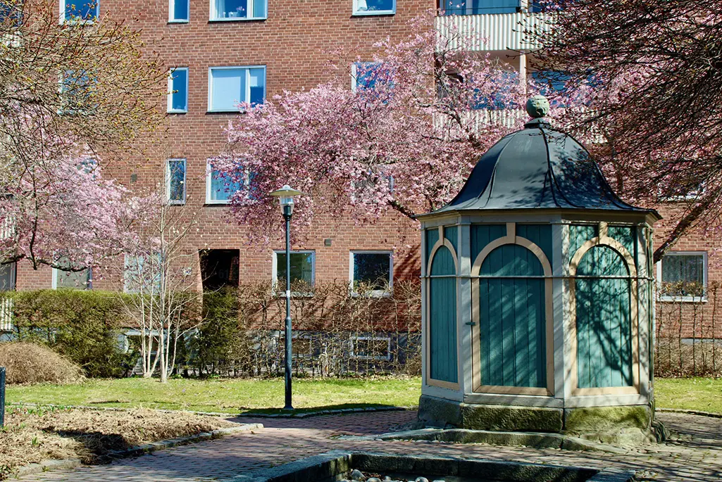 Foto på brunnshuset som står i Munksundskällan. I bilden syns även blommande körsbärsträd, buskar och gräsmatta.