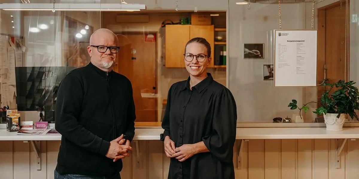En kvinna och man står i receptionen och ler välkomnande.