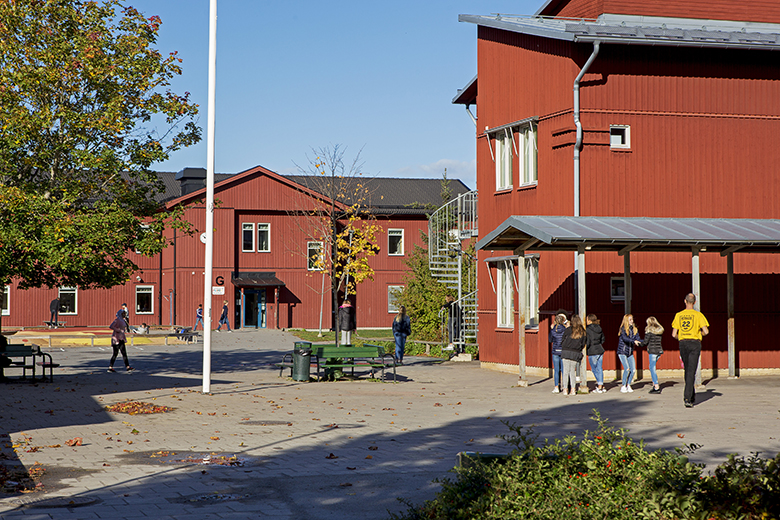 Foto på några av Örsundsbroskolans byggnader som de ser ut idag. Det är röda träbyggnader i två våningar. Mellan byggnaderna är det en asfalterad yta med några bänkar och en flaggstolpe. 