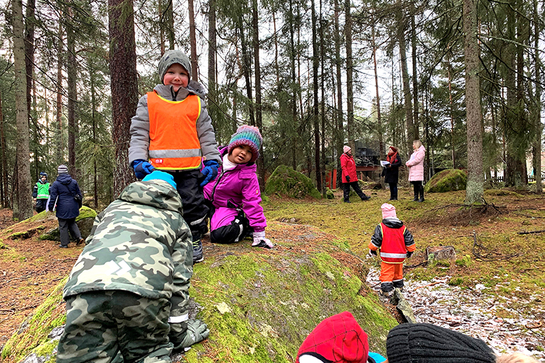 Barn klättrar på sten i skogen, andra barn och vuxna syns i bakgrunden