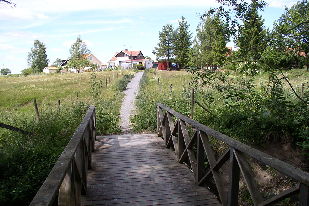Träbro som leder mot en äng på landet med hus i horisonten, foto.