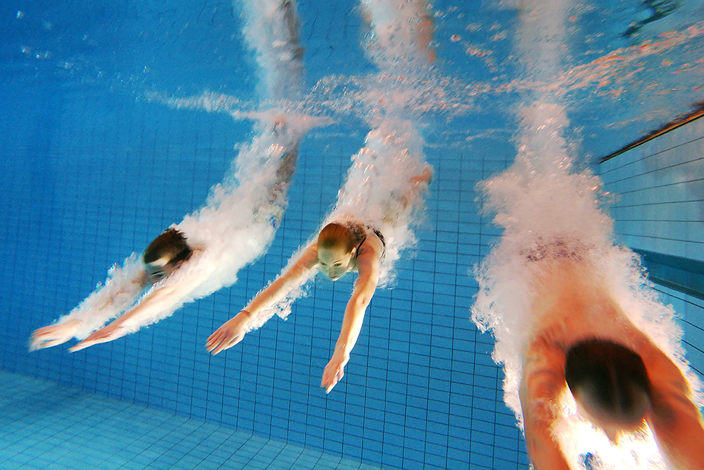 Foto på tre personer under vatten som precis dykt ner i en bassäng.