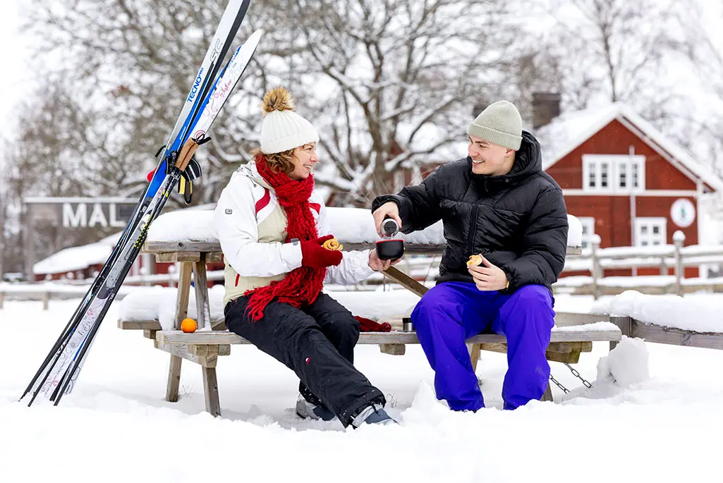 Två personer fikar utomhus i vintrig miljö med längdskidorna lutandes mot fikabordet.