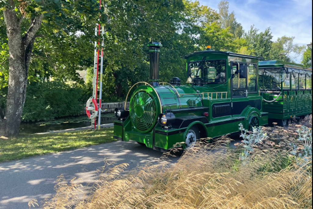 Bild på ett grönt el-tåg i parkmiljö.