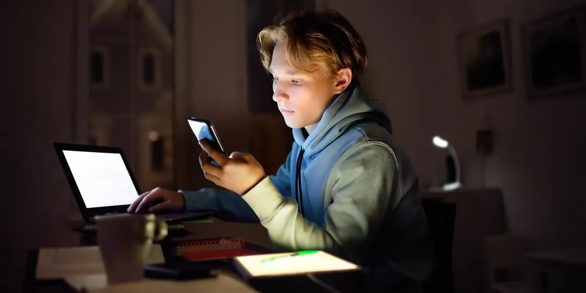 Blond ung kille som sitter framför en datorskärm och kollar på sin mobil.