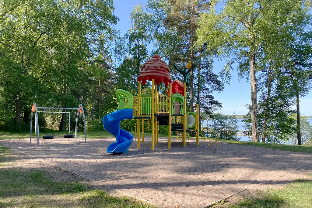 Foto på den nuvarande lekplatsen vid Bredsands bad och camping. på bilden syns en färgglad klätterställning med rutschkana samt en gungställning med två däckgungor. De står på en gruslagd yta. Klätterställningen har delar i blått, grönt, gult och rött.
