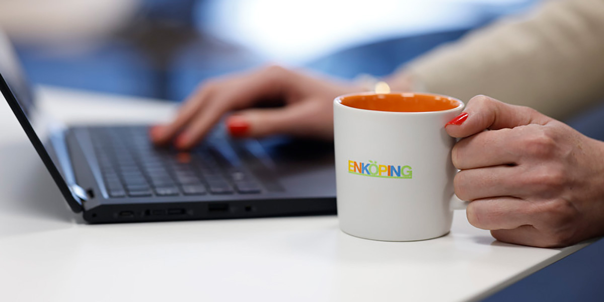 Händer som håller i kaffekopp med Enköpings logotyp och dator