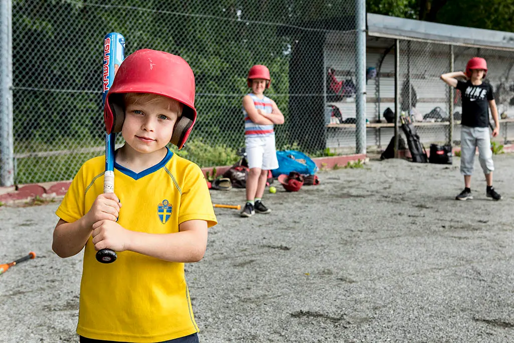 Foto på en ung pojke i svensk landslagströja. Han har en röd hjälm på sig och håller i ett basebollträ. I bakgrunden syns två andra unga pojkar med röda hjälmar på sig.