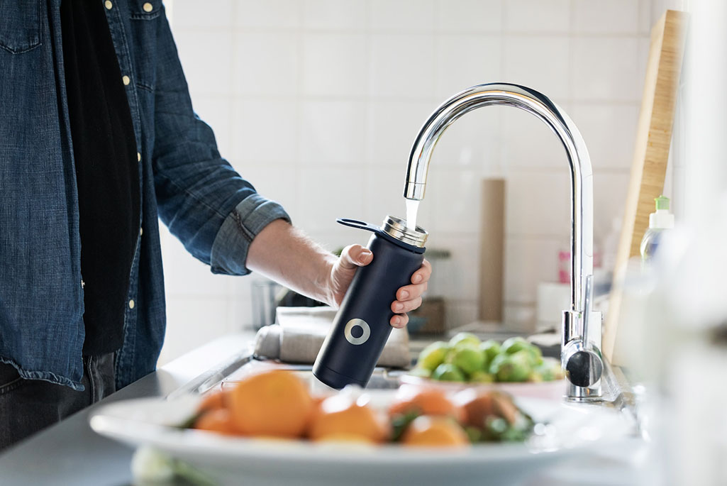 Foto på person som fyller en vattenflaska under kökskranen. I förgrunden och bakgrunden syns fat med olika frukter.