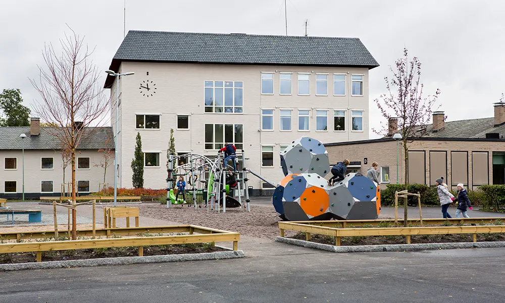 Foto på några av Korsängsskolans byggnader. Det är vita byggnader med varierande mängder våningar. I förgrunden av bilden syns skolgården med klätterställningar. 