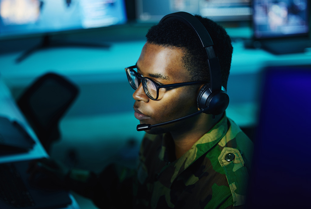 En man i militärkläder sitter med headset framför en datorskärm, foto