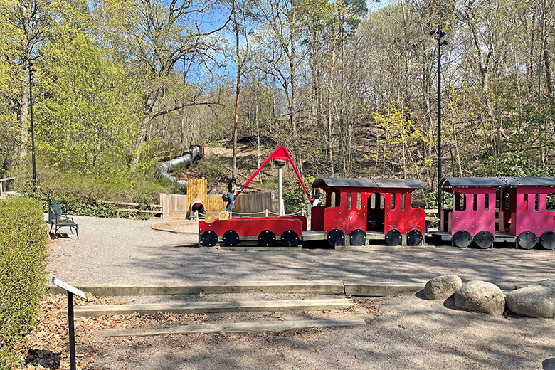 Foto på skolparkens lekplats som den ser ut idag. I bilden syns lekutrustning som ett rött litet tåg man kan sitta i. En stor gunga och en lång rutschkana som går utför en kulle in till lekplatsen.