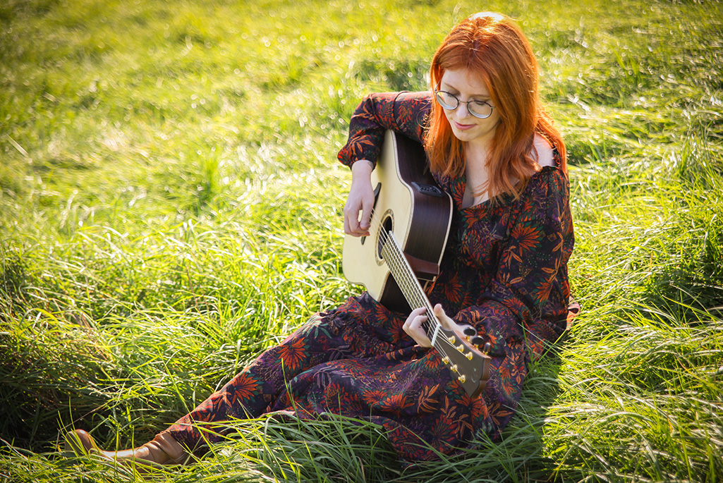 Bild på Michelle Sandberg som sitter i gräset och spelar gitarr.
