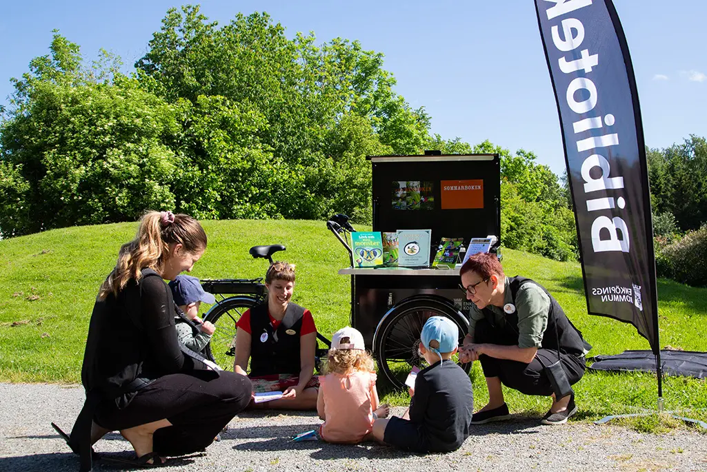 Två kvinnliga bibliotekarier sitter med en lådcykel full av böcker och visar böcker för två barn.