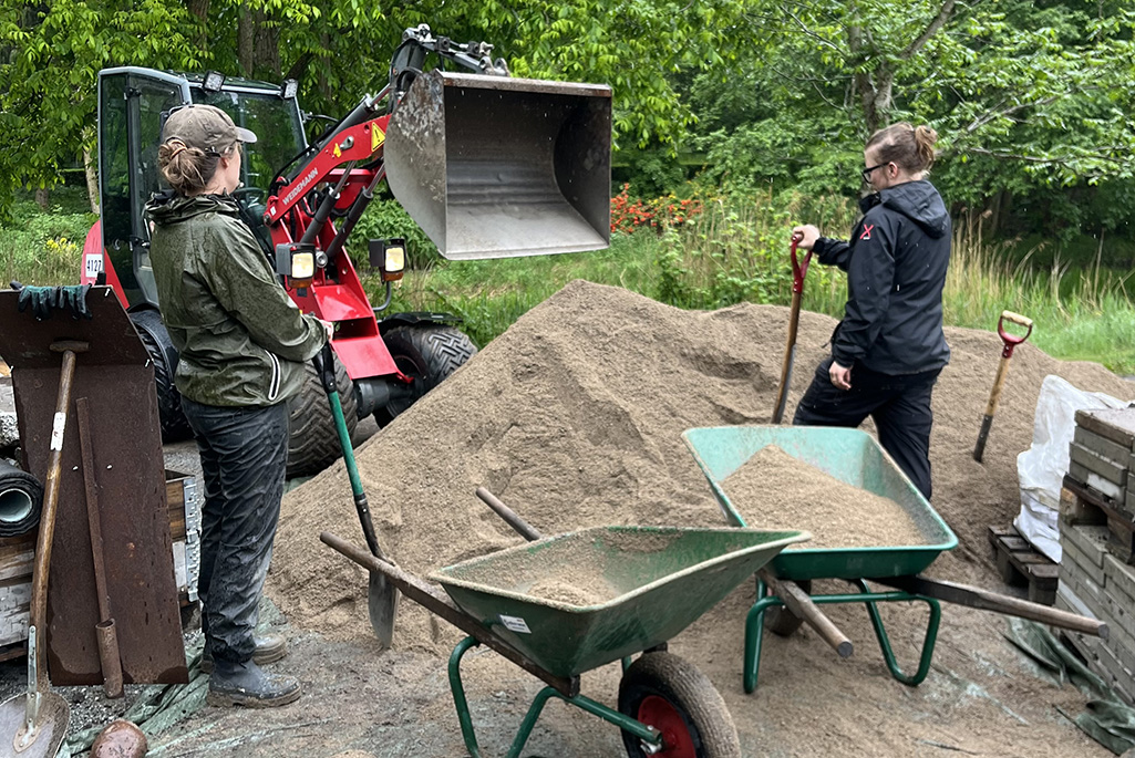 Två kvinnor vid en stor hög med sand och flera skottkärror.