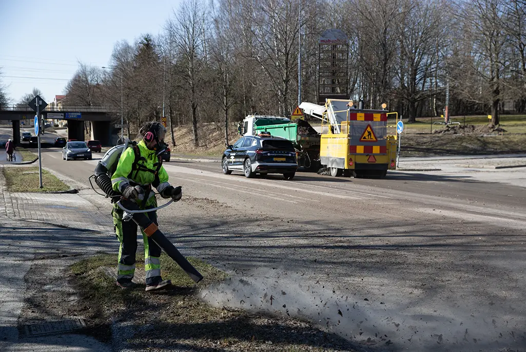 Parkarbetare i gula arbetskläder och munskydd blåser rent kanten mot en väg.